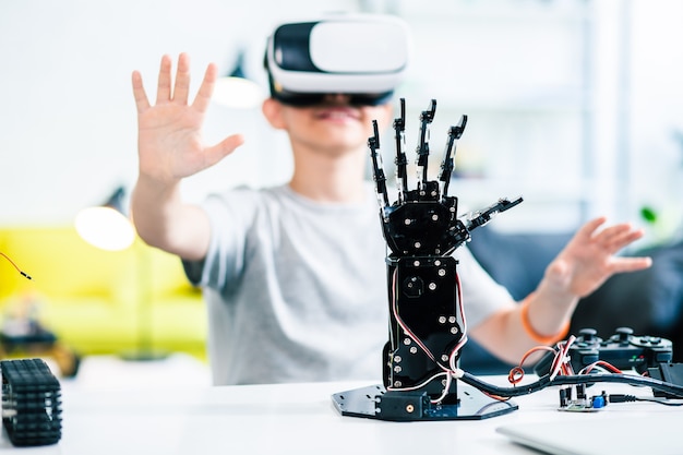 Selective focus of a robotic hand on the table with a little smiling boy testing VR glasses in the background