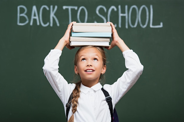 Photo selective focus of smiling schoolgirl holding stack of books above head near chalkboard with back to