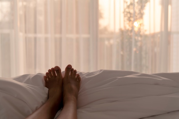 Selective focus of woman feet relax on white blanket in bedroom in the morning with warm sun light from window