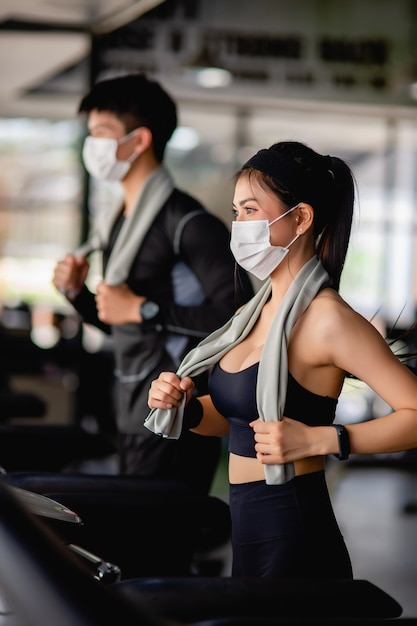 Selective focus,  young sexy woman in mask wearing sportswear and smartwatch and blurred young man. They are running on treadmill to for workout in modern gym, 