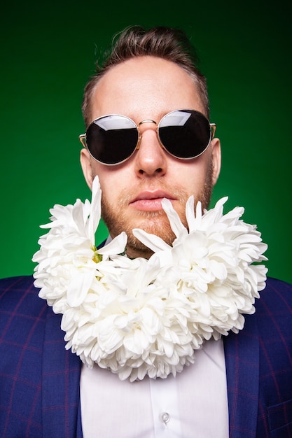 Self assured male in elegant suit and with tender white flowers in beard looking at camera on green background in studio