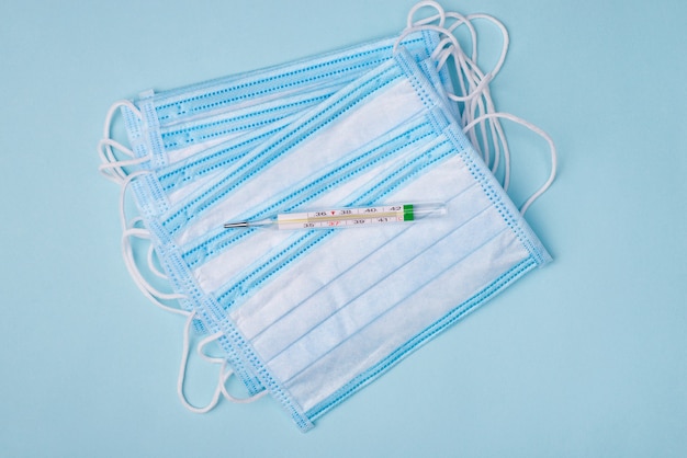 Self-isolation first symptom danger concept. Above overhead close up photo of pile of medical masks and mercury thermometer isolated on blue background