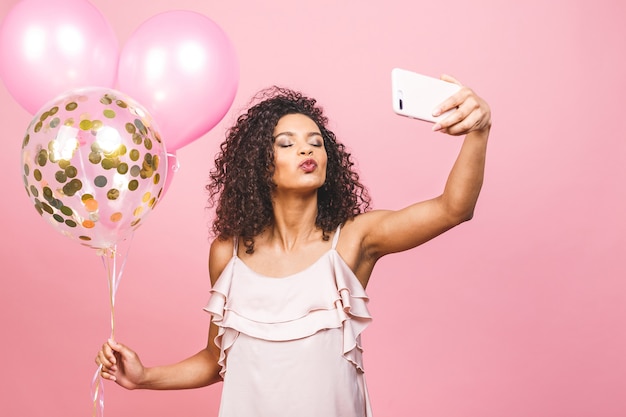 Self portrait of afro american positive girl in dress having balloons in hand shooting selfie on front camera isolated on pink background.