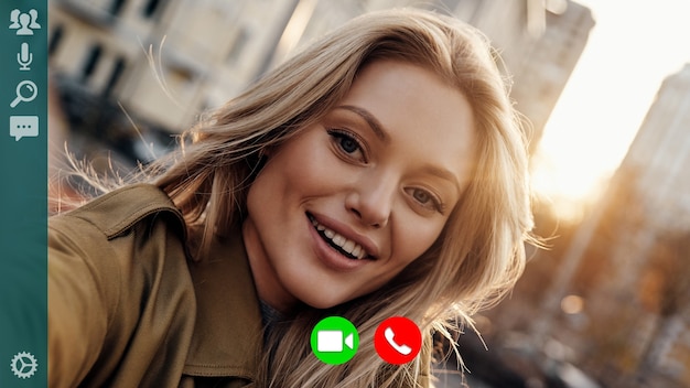 Self portrait of attractive young woman smiling and looking at camera while having video call outdoors