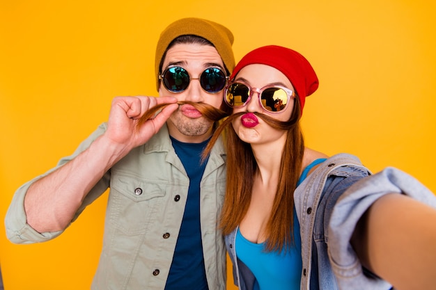 Self-portrait of   nice attractive childish humorous cheerful couple spending weekend making fake moustache having fun isolated on bright vivid shine vibrant yellow color background