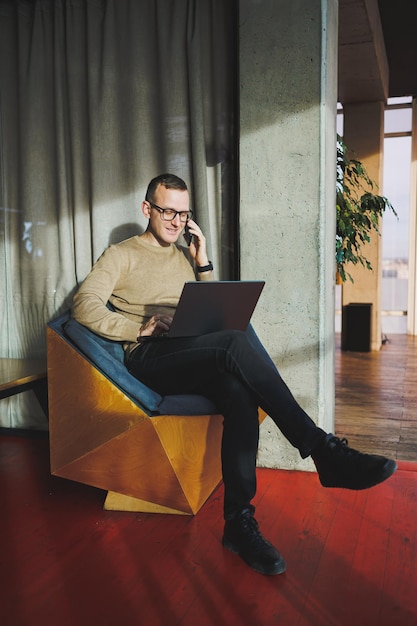 A selfemployed man in stylish clothes is making a blog website post on a laptop computer in a modern workspace Male freelancer works remotely and talks on the phone