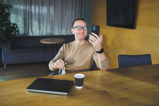 A selfemployed man in stylish clothes is making a blog website post on a laptop computer in a modern workspace Male freelancer works remotely and talks on the phone