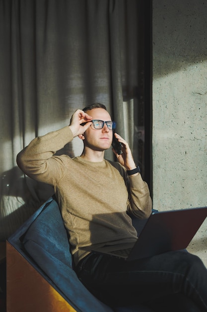 A selfemployed man in stylish clothes is making a blog website post on a laptop computer in a modern workspace Male freelancer works remotely and talks on the phone