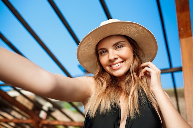 Selfie of beautiful young cheerful girl in hat and sunglasses rests at morning beach