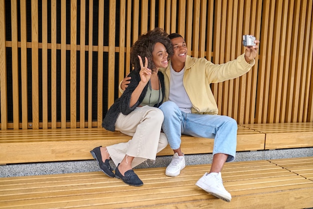 Selfie. Smiling dark-skinned guy hugging girl showing freedom sign taking selfie on camera sitting outdoors on fine day