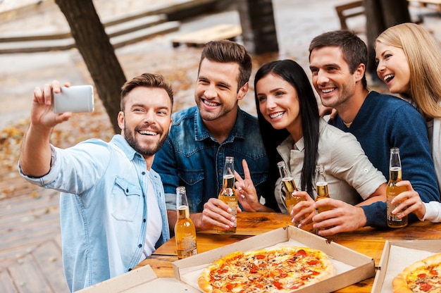 Selfie time! Group of cheerful young people bonding to each other and making selfie on smart phone while drinking beer and eating pizza together