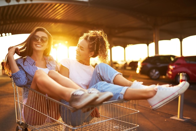 Selfie time Two beautiful girls having fun on shopping trolley Young people racing on shopping cart