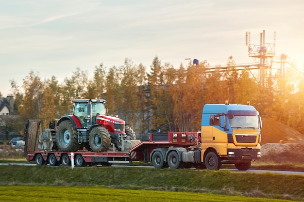 Photo the semi truck is hauling farm equipment on a flatbed the heavy industrial truck semi trailer flatbed platform transport one big modern farming tractor machine on the road evening sun