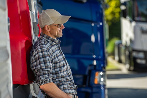 Photo semitruck driver leans against red truck in sunny transport yard during daytime