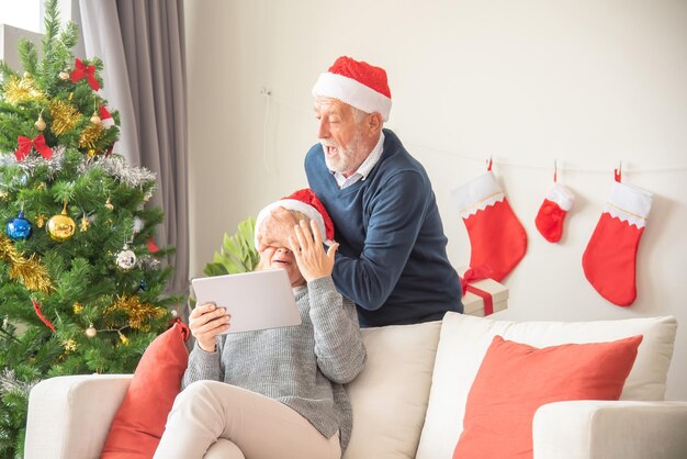 Senier husband gives a gift box to his wife While sitting on the sofa and wearing a Santa Claus hat