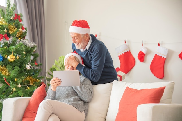 Senier husband gives a gift box to his wife While sitting on the sofa and wearing a Santa Claus hat