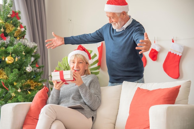 Senier husband gives a gift box to his wife While sitting on the sofa and wearing a Santa Claus hat