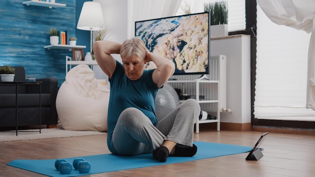 Senior adult doing physical exercise and watching video on tablet to follow online training lesson. Old woman using device to do fitness workout and gymnastics from home. Active person