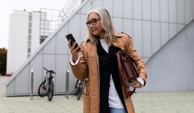 Senior adult grayhaired business woman hurries to a meeting and looks at a mobile phone year old