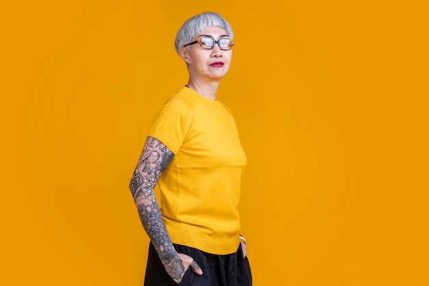 Senior asian female grey hair fashioned cloth with tattoo arm standing hand gesture with positive confident expressionold asia woman wear yellow tshirt carefree lifestyle happiness pension age