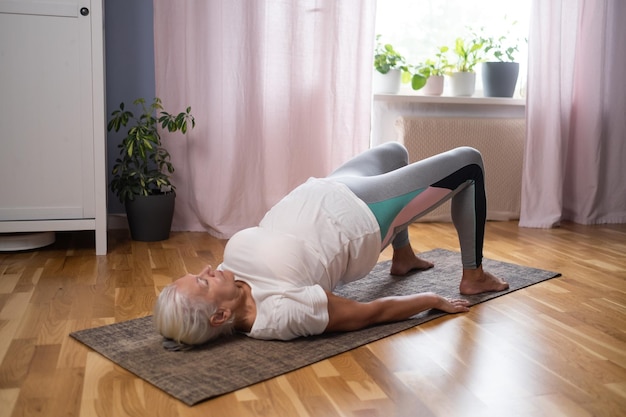 Senior athletic yoga woman doing ardha chakrasana wheel on yoga mat at home