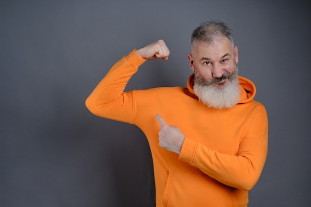 Senior bearded man dressed orange hoodie shows muscles after practising over gray studio wall, look I am very strong and healthy, perfect biceps