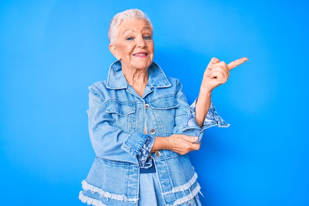 Senior beautiful woman with blue eyes and grey hair wearing casual denim style smiling happy pointing with hand and finger to the side