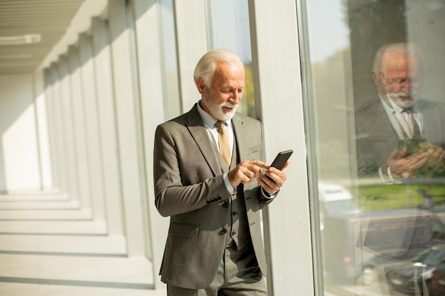 A senior business man stands in an office hallway focused on his mobile phone