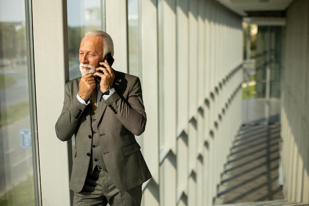 A senior business man stands in an office hallway focused on his mobile phone