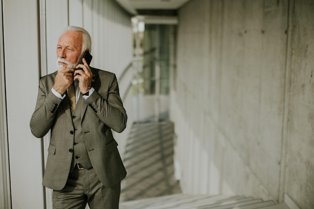 A senior business man stands in an office hallway focused on his mobile phone