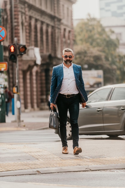 A senior businessman in a suit and sunglasses walking around the city while carrying a bag