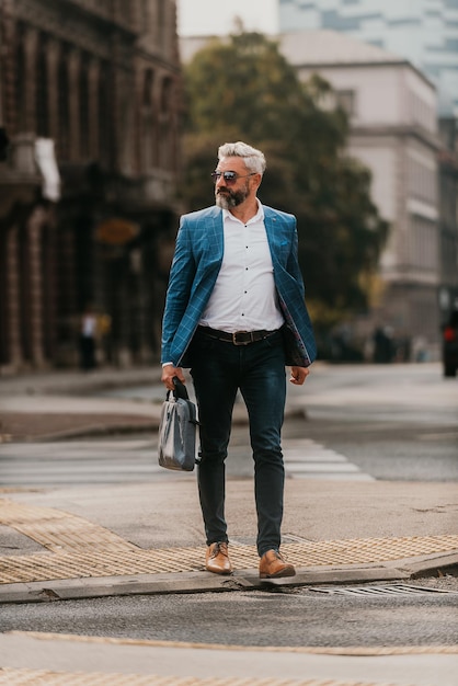 A senior businessman in a suit and sunglasses walking around the city while carrying a bag