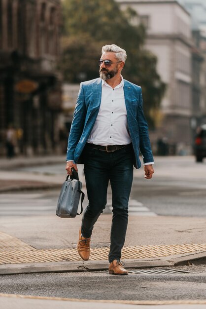 A senior businessman in a suit and sunglasses walking around the city while carrying a bag