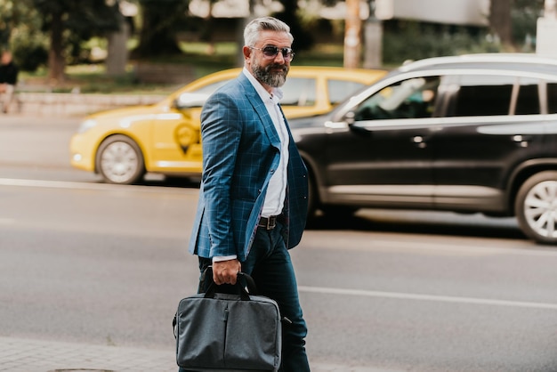 A senior businessman in a suit and sunglasses walking around the city while carrying a bag