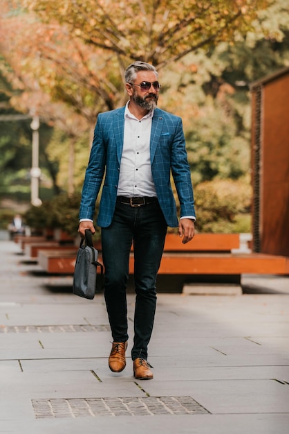 A senior businessman in a suit and sunglasses walking around the city while carrying a bag