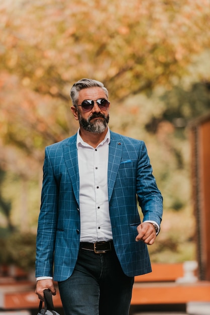 A senior businessman in a suit and sunglasses walking around the city while carrying a bag