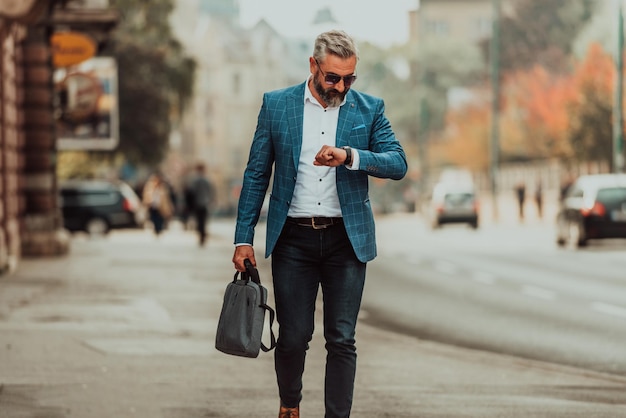 Senior businessman walking in the city carrying a bag and using a watch