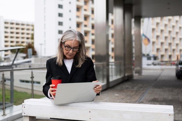 Senior businesswoman working on a laptop on the terrace of an office building with a cup of coffee