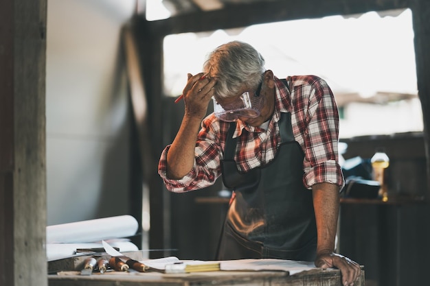 Senior carpenter use hand over head thinking or serious or sad or depression for project working on wood working machines in carpentry shop man works in a carpentry shop