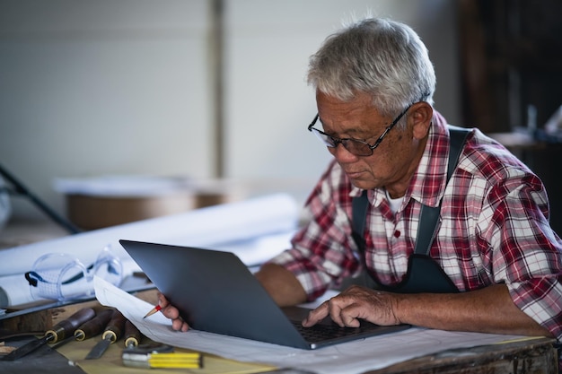 Senior carpenter using laptop work with the blueprint for project working on wood working machines in carpentry shop man works in a carpentry shop