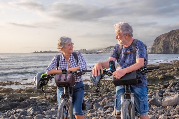 Senior caucasian couple riding on the pebble beach with bicycles at sunset looking each other smiling Authentic elderly retired life concept