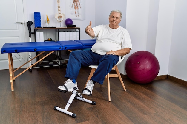 Senior caucasian man at physiotherapy clinic using pedal exerciser smiling friendly offering handshake as greeting and welcoming successful business