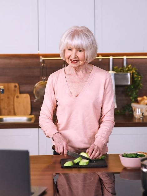 senior cheerful woman is cooking at modern kitchen by laptop