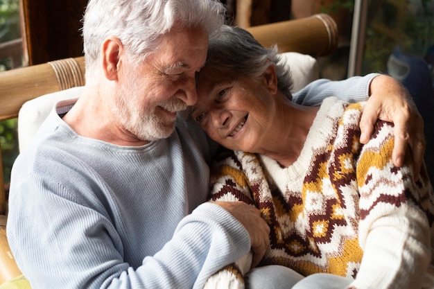 Senior couple in love and tenderness at home sitting together on the sofa. Elderly relationship lifestyle with retired man and woman smiling and having care each other