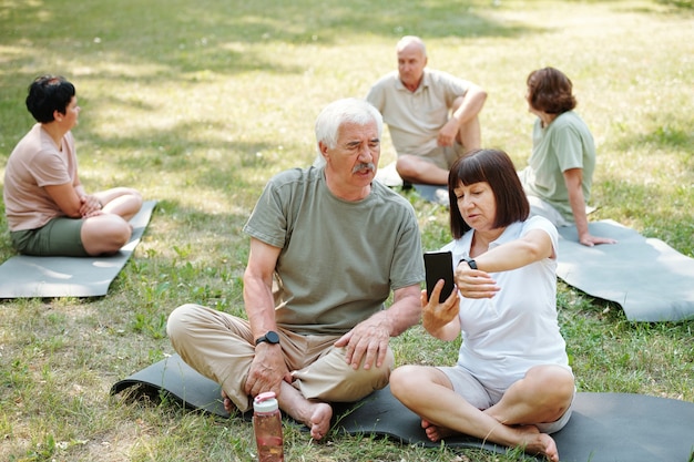 Senior couple sitting on the grass and discussing sports tracker on their gadgets during sports training in the park