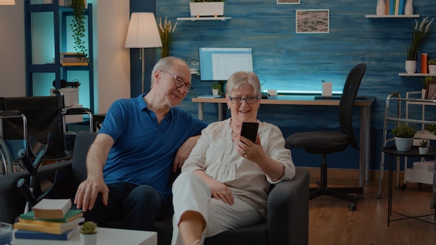 Senior couple talking to relatives on online video call at home, using smartphone to chat. Retired people with chronic disability holding phone to have remote conversation on teleconference.