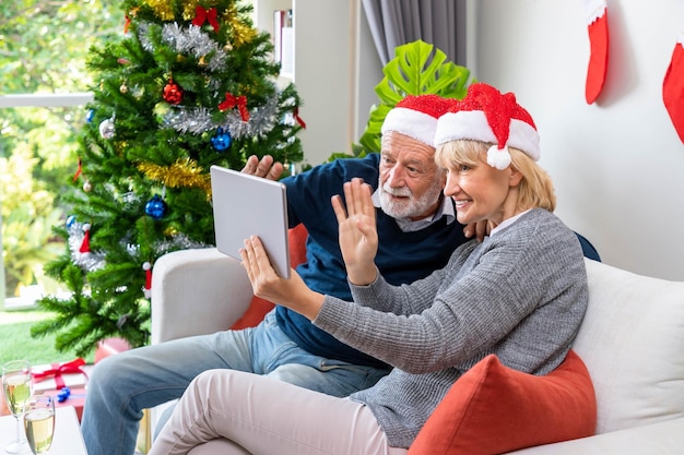 Senior couple using tablet to video phone call to greeting their family for Christmas festival wave hand sitting on sofa with decoration and tree