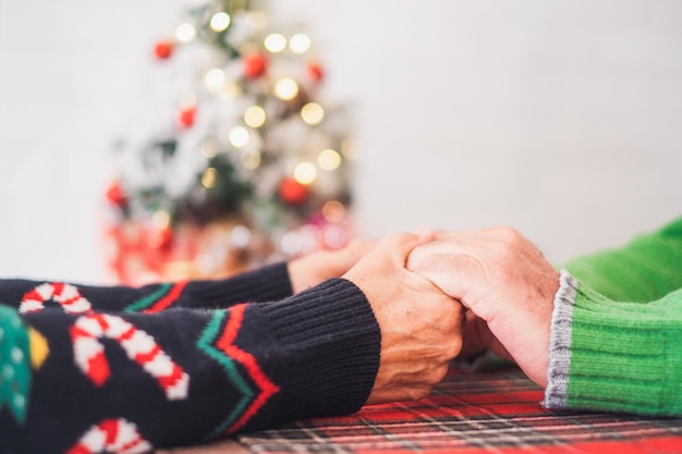 Senior couple in warm clothing holding each others hands in front of decorated christmas tree at home Loving old romantic heterosexual couple celebrating christmas festival togetherxA