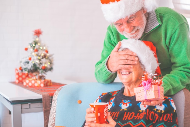 Senior couple in warm clothing and santa hat man covering eyes of old wife with a surprise gift for her Loving old romantic heterosexual couple celebrating christmas festival togetherxA