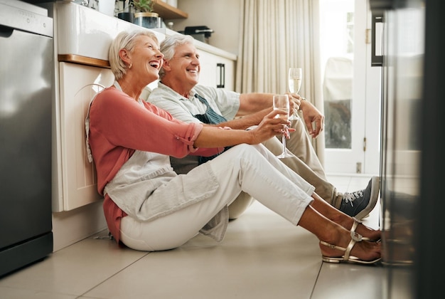 Senior couple wine and laughing on kitchen floor thinking happy memories and talking about love during a celebration of anniversary new home or retirement Old man and woman telling a funny joke